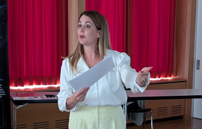 Tamara steht mit einem Blatt in der Hand vor rotem Vorhang und spricht zum Publikum